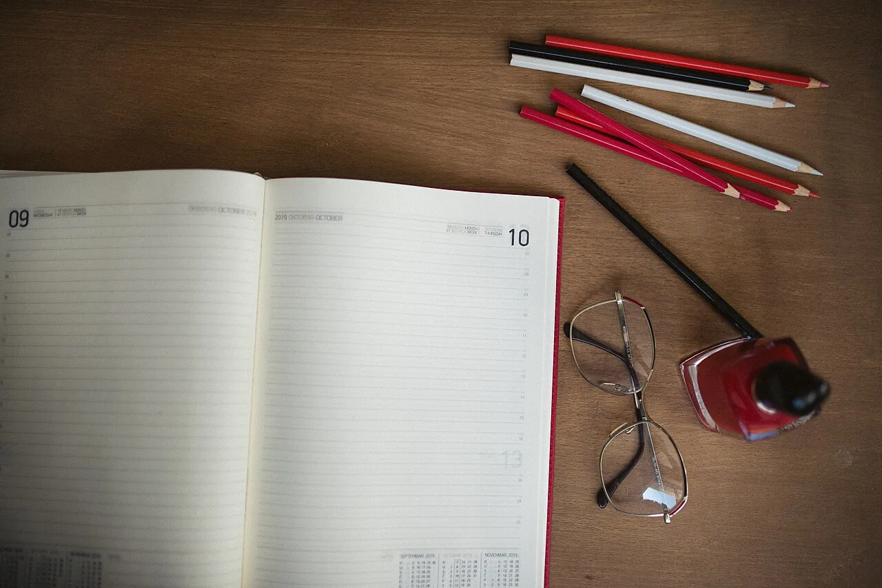 An open planner, pencils, glasses, and a drink on a wooden desk