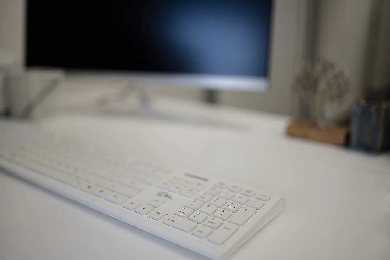 A white keyboard on a desk with a monitor in the background