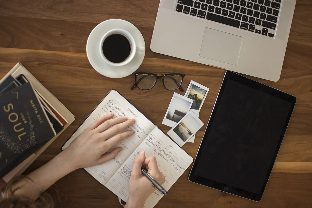 Hands writing in a notebook beside a laptop and coffee