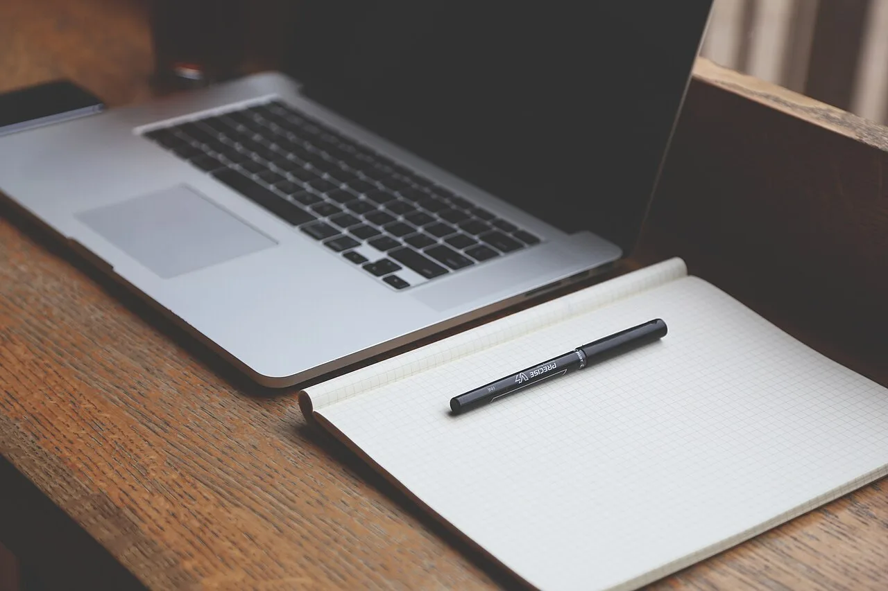 A laptop, blank notebook, and pen on a wooden desk
