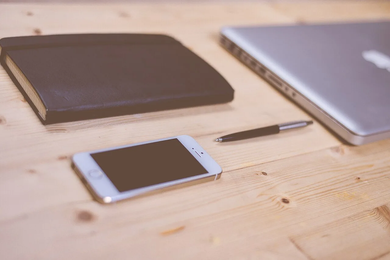 A phone, notebook, pen, and laptop arranged on a pale wooden desk