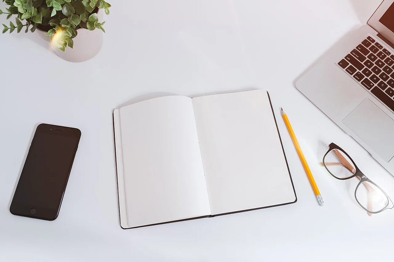 A blank notebook, phone, pencils, and laptop on a white desk