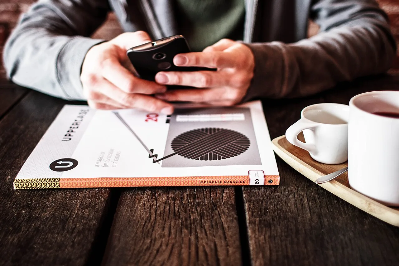 Hands holding a phone beside a coffee cup and reading material